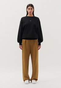 Felpa nera con maniche raglan e dettaglio del logo, abbinata a pantaloni giallo senape a vita alta e sneakers bianche su uno sfondo neutro.