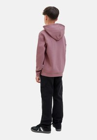 Enfant de dos, portant un sweat à capuche rose et un pantalon cargo noir, avec des baskets noires. L'arrière-plan blanc met en valeur la tenue.
