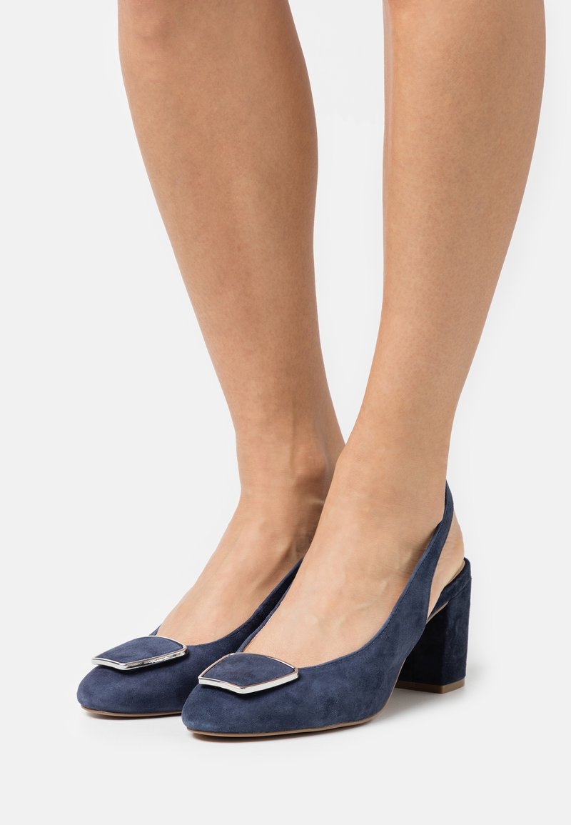 Jambes portant des escarpins slingback en daim bleu à talons blocs avec des boucles décoratives métalliques carrées sur les orteils.