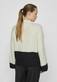 Stickad tröja med en delad design; krämfärgad överdel med ribbad textur och svart underdel med slät yta. Långa ärmar.
