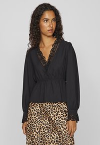 Musta pitkähiha blouse syvällä V-kaula-aukolla, pitsireunalla ja kerrotulla vyötäröllä. Puvun päällä on leopardikuosiset hameet, joissa on teksturoitu kangas ja pitsireunukset.