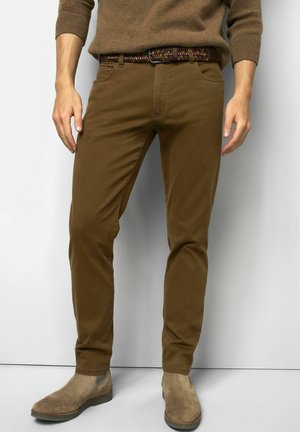 Man wearing brown slim-fit pants, beige suede boots, a brown sweater, and a multicolored woven belt standing against a white wall.