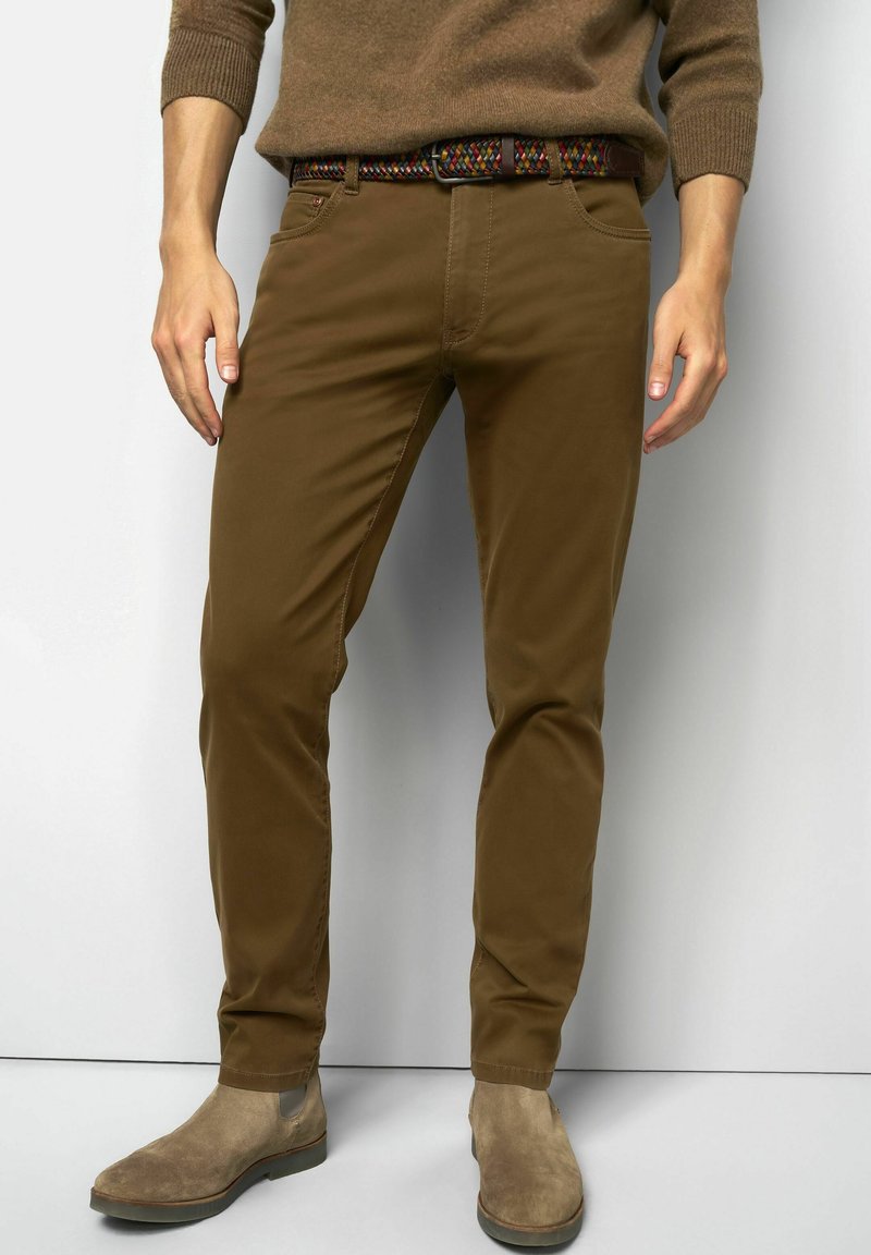 Mann in braunen Slim-Fit-Hosen, beigen Wildlederstiefeln, einem braunen Pullover und einem mehrfarbigen gewebten Gürtel, der an einer weißen Wand steht.