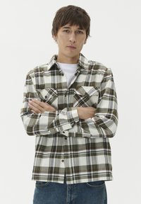 Młody mężczyzna z założonymi rękami, ubrany w koszulę w białą, szarozieloną kratę na białym podkoszulku oraz niebieskie dżinsy, na tle jednolitego białego tła.