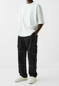 Vit bomullsstor t-shirt i oversized modell, kombinerad med svarta cargobyxor med kontrastsömmar och flera sidofickor, avslutad med vita sneakers.