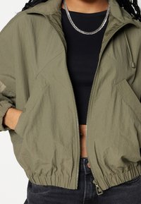 Person wears olive green zip-up jacket over black cropped top, paired with dark jeans and silver chain necklace, hand in jacket pocket.