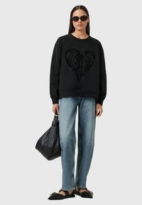 Felpa nera con un design a cuore testurizzato, abbinata a jeans blu chiaro e accessori neri, tra cui una borsa e scarpe.