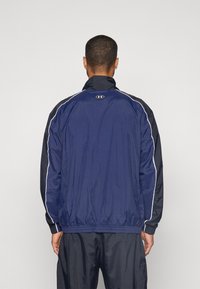 Mørkeblå sportsjakke med sorte detaljer, elastisk talje og hvide kanter. Har en høj krave og et materiale med en glat tekstur.
