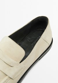 Beige suede loafer with a smooth black leather interior, featuring a wide strap across the upper and a rounded toe. Flat black sole.
