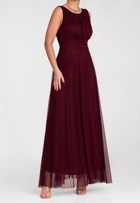 Femme portant une robe de soirée bordeaux sans manches, longue jusqu'au sol, avec une taille cintrée et une jupe fluide, associée à des talons ouverts couleur nude.