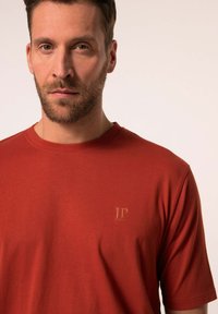 Červené bavlněné tričko s kulatým výstřihem, krátkými rukávy a malým embosovaným logem na hrudi. Hladká textura s přiléhavým střihem.