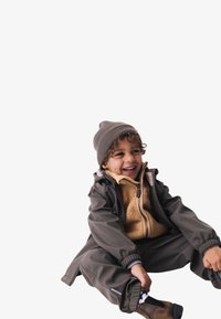 Ein Kind trägt eine dunkelgraue wasserdichte Jacke über einem beigen Fleece, kombiniert mit passenden Hosen und einer grauen Strickmütze, und sitzt vor einem weißen Hintergrund.