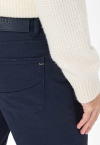 Navy blauwe textuur broek met een achterzak, voorzien van een klein merkembleem. De drager heeft een witte gebreide trui met ribgebreide manchetten.