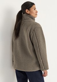 Harmaa fleece-takki, jossa on korkea kaulus ja teksturoitu pinta. Takissa on rento malli ja ommeltu yksityiskohta hihoissa.