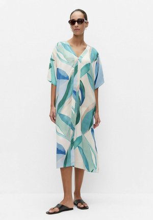 Frau trägt ein lockeres, knielanges, geknöpftes Kleid mit blauen und grünen abstrakten Mustern, schwarze Sandalen, dunkle Sonnenbrille und Creolen.