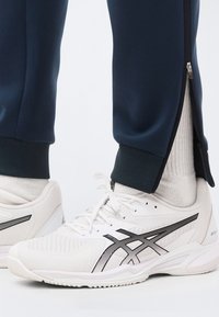 Λευκά αθλητικά παπούτσια με μαύρες ρίγες, φορεμένα με λευκές κάλτσες και ναυτικού χρώματος παντελόνια με ανοιχτά φερμουάρ στους αστραγάλους.