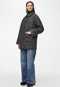 Cappotto grigio in misto lana con colletto ampio, bottoni frontali e due tasche laterali. Abbinato a jeans in denim blu larghi e calzature nere.