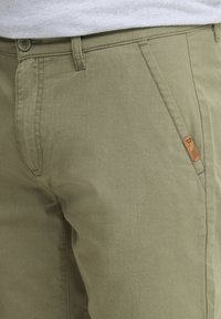 Khakihosen aus weichem Baumwollstoff, mit glatter Textur, schrägen Seitentaschen und einem kleinen braunen Logosticker nahe dem Bund.