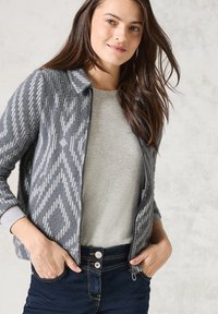 Cardigan gris avec un motif en chevrons, présentant une fermeture éclair à l'avant et des poignets côtelés, superposé sur un haut gris clair et un jean en denim foncé.