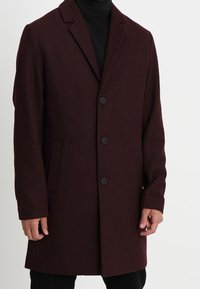 Cappotto in misto lana bordeaux con rever a cresta, quattro bottoni neri, tasche laterali e vestibilità strutturata, indossato sopra un dolcevita nero.