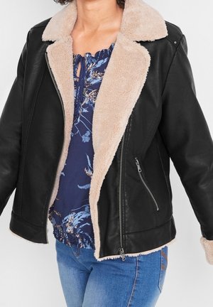 Femme portant une veste en cuir noire avec une doublure en peau lainée beige, un haut bleu à motifs floraux et un jean bleu sur un fond uni.