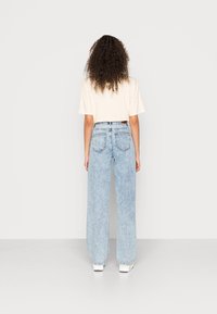 Jeans de perna larga em azul claro, feitos de denim, com uma cintura alta, dois bolsos traseiros e detalhes de costura sutis. Combinados com uma t-shirt creme.
