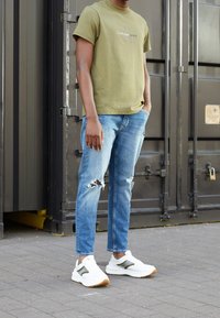 Olivgrön bomullströja med logotyp, slitna ljusblå jeans och vita sneakers med gröna detaljer, stående på grått asfalt.