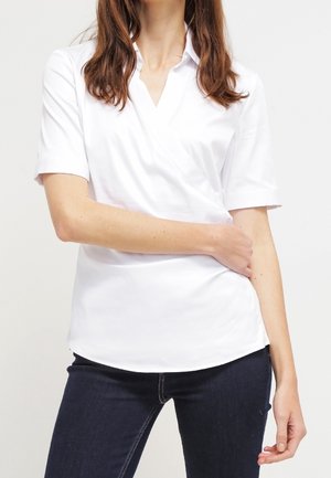 Blouse blanche à manches courtes en portefeuille avec un col en V, une texture lisse et une coupe décontractée, associée à un jean bleu foncé.