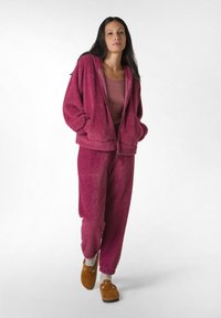 Felpa rosa fuzzy con zip e pantaloni abbinati realizzati in materiale morbido, abbinati a pantofole marroni e calzini grigi. Design accogliente e vestibilità rilassata.