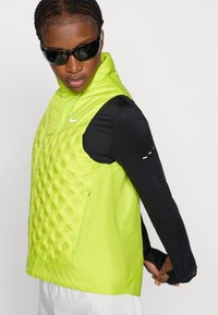 Λαμπερό πράσινο γιλέκο με επένδυση, υψηλό γιακά και τσέπες με φερμουάρ, συνδυασμένο με μαύρη μακρυμάνικη μπλούζα με αντανακλαστικές λεπτομέρειες.