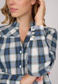 Blouse à carreaux bleu et gris avec un col haut, des manches bouffantes et trois boutons décoratifs au niveau des poignets. Fabriquée en tissu léger.