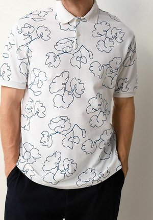 Biała koszulka polo z krótkim rękawem i czarnym konturem wzoru kwiatowego, noszona przez osobę z rękami w kieszeniach ciemnych spodni.