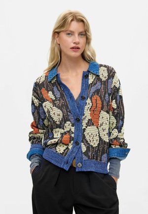 Donna bionda che indossa un cardigan multicolore a motivi con colletto e polsini in denim blu, pantaloni neri e mani nelle tasche su sfondo bianco.