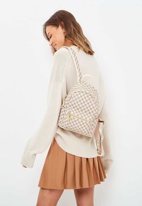 Kraftigt mönstrad ryggsäck i bruna nyanser med gyllene dragkedjor, med en rundad design och justerbara axelremmar. Bärs över en krämfärgad tröja och en plisserad kjol.