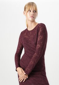 Jeune femme aux cheveux blonds en queue de cheval, portant une robe en dentelle bordeaux ajustée à manches longues, regardant sur le côté avec les mains jointes devant.