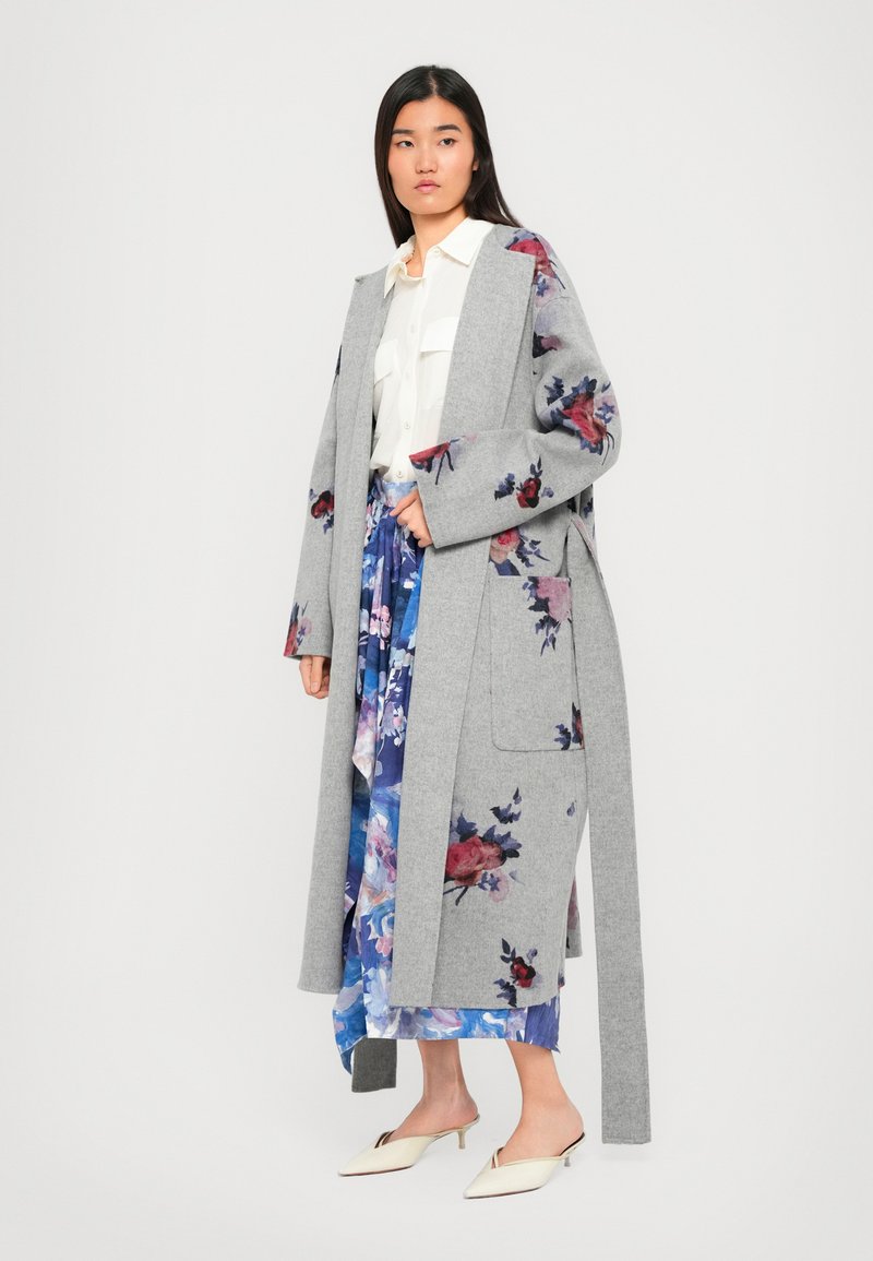 Donna con un lungo cappotto floreale grigio indossato su una camicia bianca e una gonna floreale blu, abbinato a delle mules con tacco kitten a punta color crema.