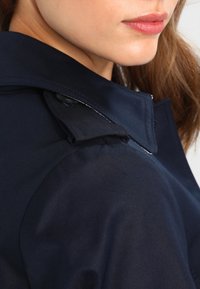 Mörkblå trenchcoat med en strukturerad krage, texturerat tyg, subtil glans och knapprad på axeln.