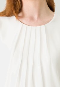 Blouse blanche avec un décolleté arrondi et des plis verticaux, fabriquée à partir d'un tissu léger et lisse au toucher doux.