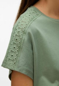 Mėtų žalios spalvos marškinėliai trumpomis rankovėmis su detaliu nėriniu iškirptėje, lygus medvilnės audinys ir laisvas fasonas.