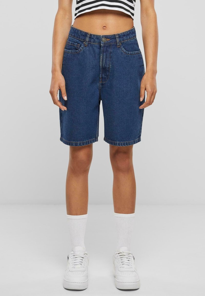 Personne portant un short en jean bleu foncé taille haute, des chaussettes blanches, des baskets blanches et un top court rayé noir et blanc, debout sur un fond gris.