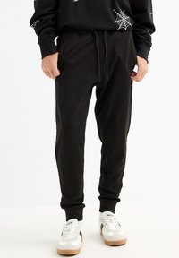Pantalones de chándal negros, hechos de una suave mezcla de algodón, que cuentan con una cintura con cordón, piernas ajustadas y bolsillos laterales. Sin patrones ni adornos.