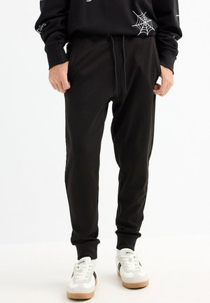 Pantalones de chándal negros, hechos de una suave mezcla de algodón, que cuentan con una cintura con cordón, piernas ajustadas y bolsillos laterales. Sin patrones ni adornos.