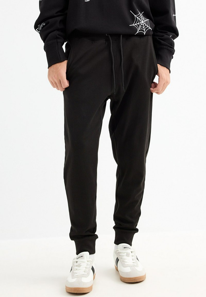 Pantalones de chándal negros, hechos de una suave mezcla de algodón, que cuentan con una cintura con cordón, piernas ajustadas y bolsillos laterales. Sin patrones ni adornos.