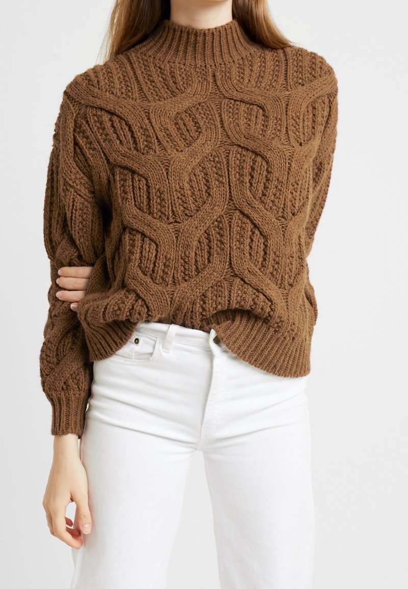 Bruine gebreide trui met een hoge hals en een textuur kabelpatroon, met lange mouwen en een iets losse pasvorm, gecombineerd met witte jeans.
