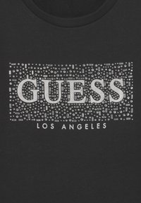 Černé tričko s stříbrným logem "GUESS" s texturovaným vzorem malých hřebíčků a obdélníků, pod nímž je označení "LOS ANGELES".