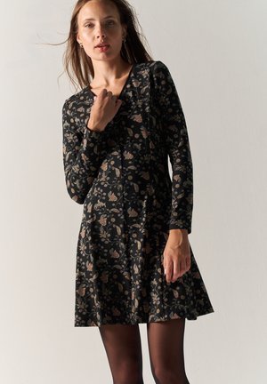 Mujer vestida con un vestido negro de manga larga con un patrón floral, de pie contra un fondo claro y liso, mirando a la cámara.