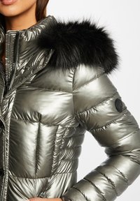 Metalická šedá bunda s výplní a černou umělou kožešinou na kapuci. Obsahuje prošívaný design, zipové zapínání a malou nášivku s logem na rukávu.