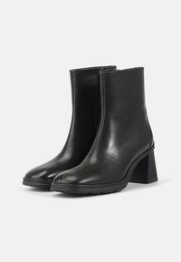 Botas de tobillo de cuero negro con una textura suave, punta cuadrada y un tacón grueso. Cremallera lateral para facilitar su uso. Diseño minimalista moderno.