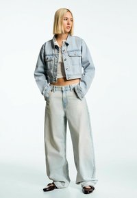 Giacca di jeans cropped azzurra chiara con chiusura a bottone e due tasche frontali, abbinata a jeans a gamba larga azzurri chiari. Sotto una maglietta bianca semplice.