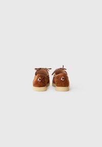 Brown suede shoes with a soft texture, featuring a lace-up design and a silver accent logo on the back. Flat rubber sole.